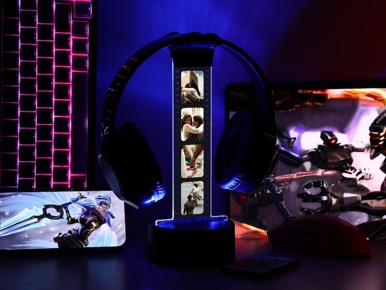 May include: A black headset rests on a lighted headphone stand displaying photos. A keyboard and a smartphone with a game graphic are visible. The scene is illuminated with blue and pink lights, creating a gaming setup atmosphere.