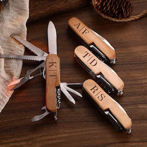 May include: A collection of personalized wooden pocket knives with various tools, including a blade, scissors, and saw. Each knife has initials engraved on the wooden handle. The knives are displayed on a dark wooden surface.