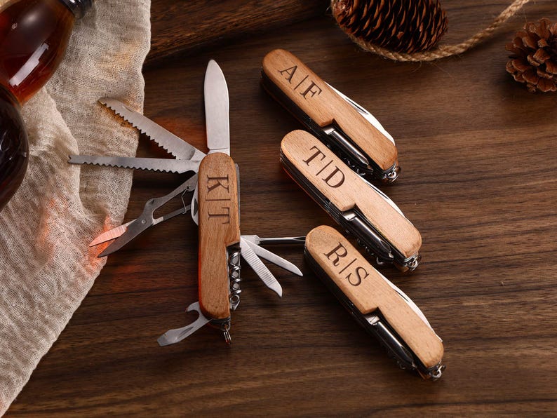 May include: A collection of personalized wooden pocket knives with various tools, including a blade, scissors, and saw. Each knife has initials engraved on the wooden handle. The knives are displayed on a dark wooden surface.
