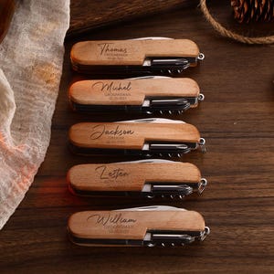 May include: Five personalized wooden multi-tools with names engraved on the wooden handle. Each tool includes a knife, screwdriver, and corkscrew. The names are Thomas, Michel, Jackson, Zesten, and William. The tools are on a dark wooden surface.