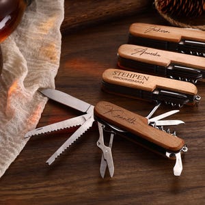 Könnte beinhalten: Eine Sammlung personalisierter Holz-Multitools mit verschiedenen Werkzeugen, darunter Messer, Korkenzieher und Scheren. Die Werkzeuge sind mit Namen und dem Wort "Groomsman" graviert. Die Werkzeuge liegen auf einer dunklen Holzoberfläche.