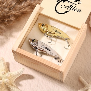 May include: A wooden box with a hinged lid, containing two personalized fishing lures. One lure is gold-coloured and the other is silver. The lid is engraved with a fish graphic and the name "Allen". The lures are engraved with names.