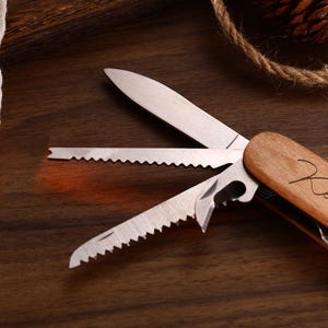 May include: A multi-tool with a wooden handle and stainless steel blades. The blades include a knife, saw, and bottle opener. The handle is engraved with the name "Kenneth". The tool is displayed on a dark wood surface.