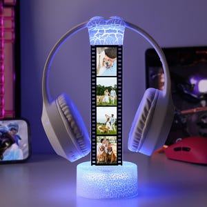 May include: A white wireless headset with a film strip of photos in the center, illuminated by blue and white lights. The base is a glowing cylinder. A computer keyboard, phone, and mouse are in the background.