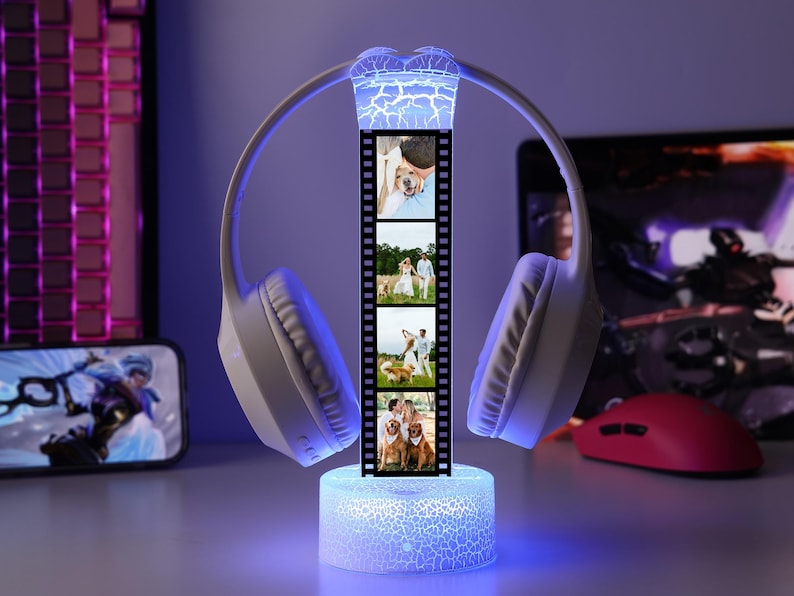 May include: A white wireless headset with a film strip of photos in the center, illuminated by blue and white lights. The base is a glowing cylinder. A computer keyboard, phone, and mouse are in the background.