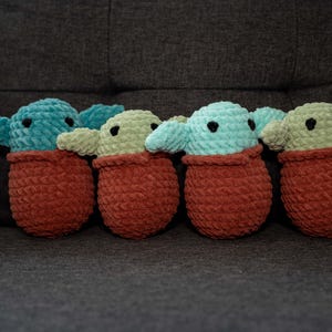 May include: Five crocheted plush toys in the shape of a popular fictional character. The toys are green and blue and are wearing brown crocheted outfits.