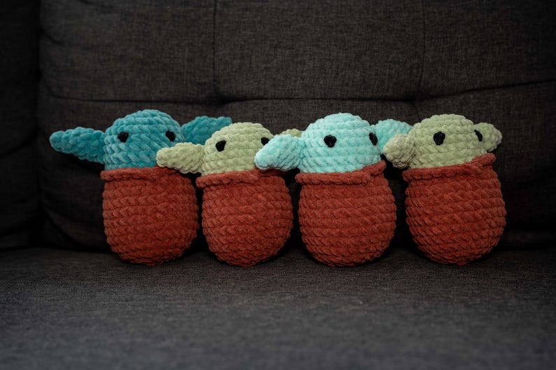 May include: Five crocheted plush toys in the shape of a popular fictional character. The toys are green and blue and are wearing brown crocheted outfits.