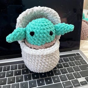 Puede incluir: Un juguete de peluche tejido a crochet de un personaje verde con ojos grandes, sentado dentro de una cesta blanca tejida a crochet.