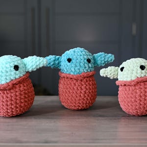 May include: Three crocheted plush toys in the shape of a popular fictional character. The toys are all wearing a red crocheted blanket. The toys are green, blue, and light green.