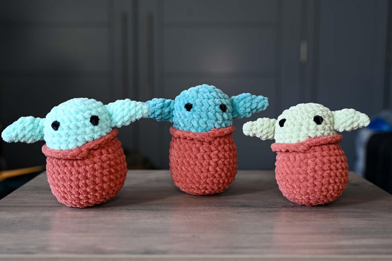 May include: Three crocheted plush toys in the shape of a popular fictional character. The toys are all wearing a red crocheted blanket. The toys are green, blue, and light green.
