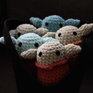 May include: A group of five crocheted plush toys in a black fabric bag. The toys are in various shades of blue and gray, with one toy having a red crocheted collar.
