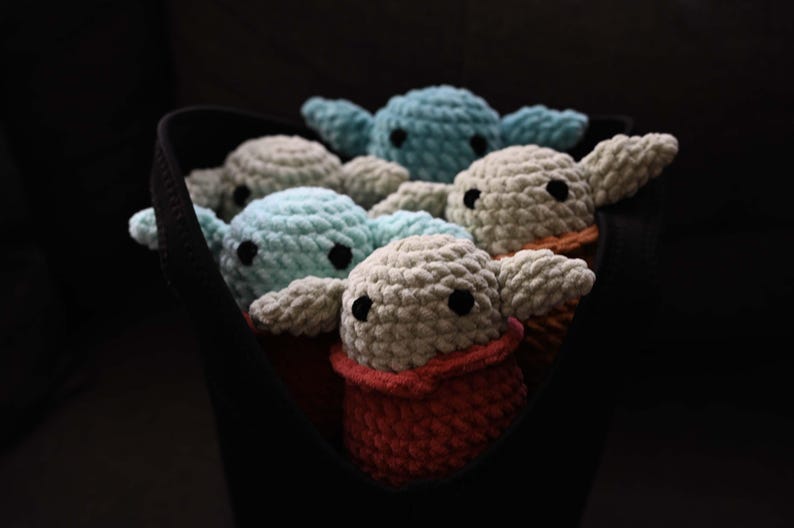 May include: A group of five crocheted plush toys in a black fabric bag. The toys are in various shades of blue and gray, with one toy having a red crocheted collar.
