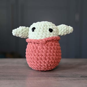 May include: A crocheted plush toy in the shape of a character from the Star Wars franchise. The toy is green and has large, black eyes. It is wearing a pink crocheted outfit.