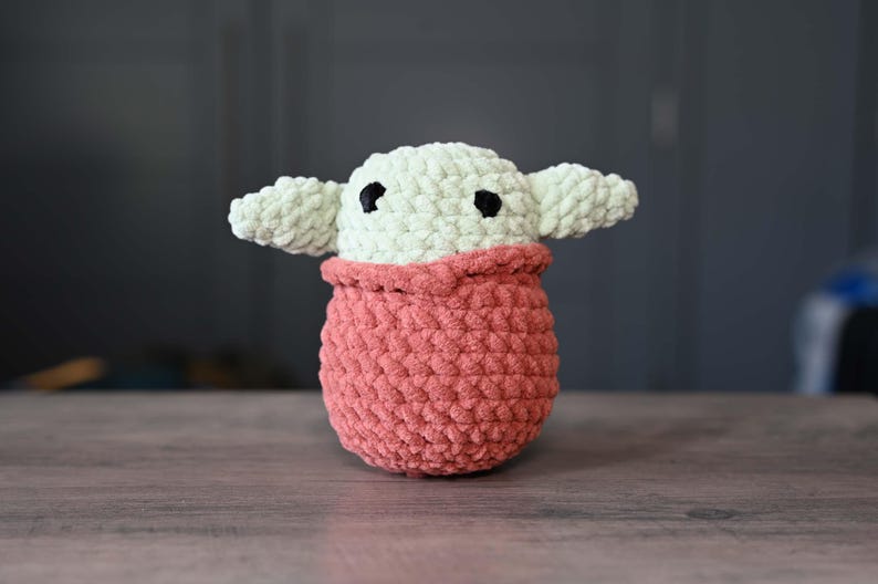 May include: A crocheted plush toy in the shape of a character from the Star Wars franchise. The toy is green and has large, black eyes. It is wearing a pink crocheted outfit.