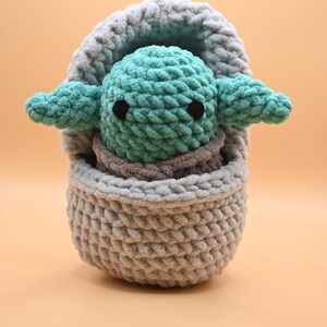 Puede incluir: Un juguete de peluche tejido a crochet de un personaje verde de la franquicia Star Wars, sentado en una cesta de crochet gris.