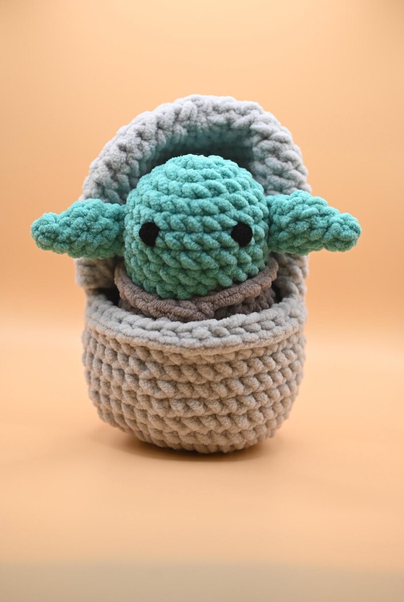 Puede incluir: Un juguete de peluche tejido a crochet de un personaje verde de la franquicia Star Wars, sentado en una cesta de crochet gris.