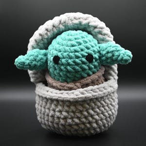 Puede incluir: Un juguete de peluche tejido a crochet de un personaje verde con ojos grandes, sentado en una cesta tejida a crochet gris y blanca.