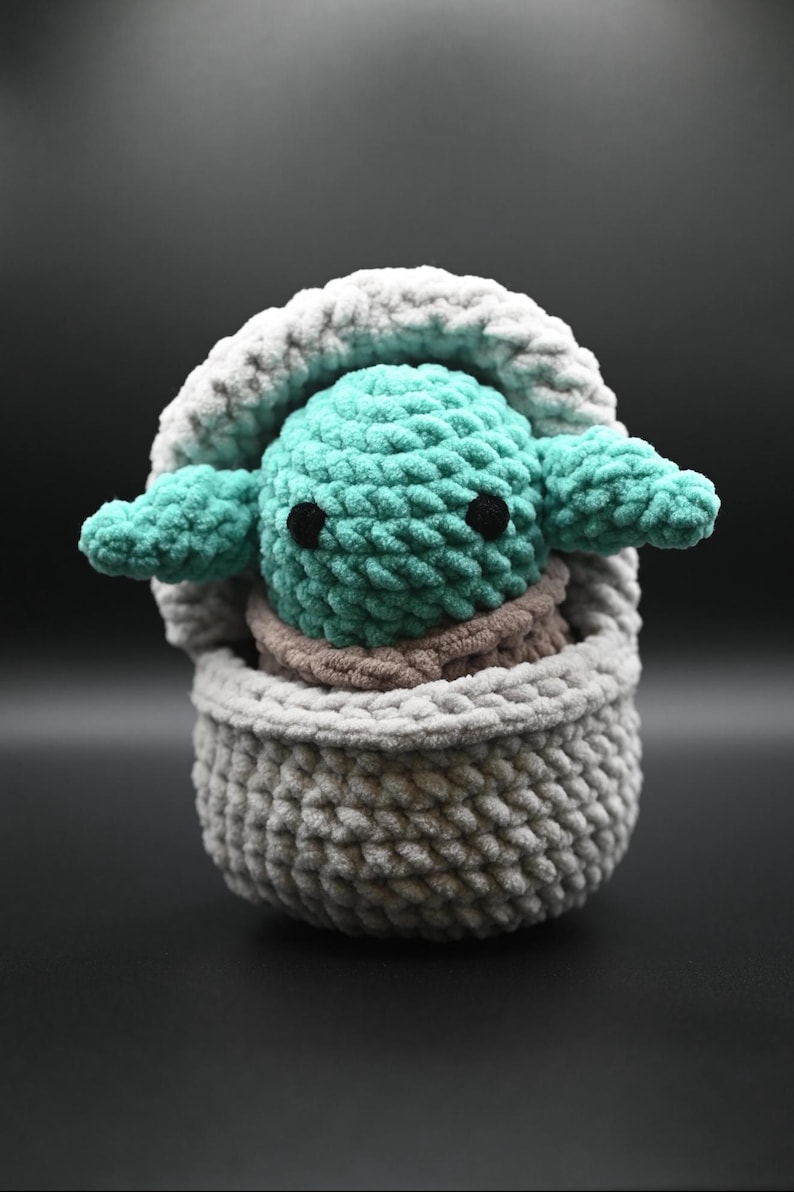 Puede incluir: Un juguete de peluche tejido a crochet de un personaje verde con ojos grandes, sentado en una cesta tejida a crochet gris y blanca.