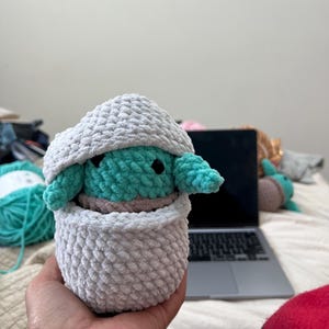 Puede incluir: Un juguete de peluche tejido a crochet con forma de personaje alien&iacute;gena verde, con ojos grandes y una peque&ntilde;a boca, est&aacute; sentado dentro de una cesta blanca tejida a crochet. La cesta est&aacute; sentada en la mano de una persona.