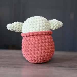 May include: A crocheted plush toy shaped like a character from the Star Wars franchise. The toy is green and pink and has large ears.