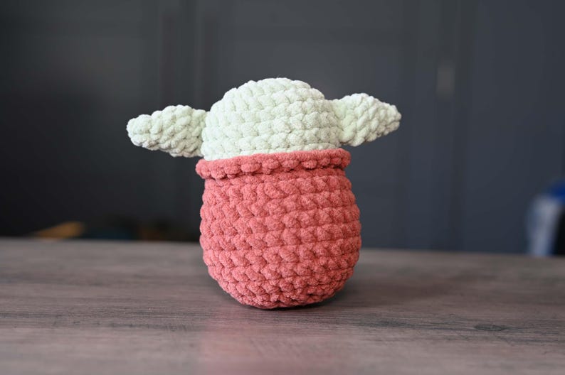 May include: A crocheted plush toy shaped like a character from the Star Wars franchise. The toy is green and pink and has large ears.