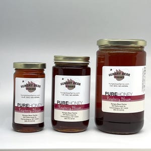 May include: Three glass jars of honey with gold lids. The jars are labeled "Hungry Bear Farms" and "Pure Honey Raspberry Blossom". The jars are different sizes, with the largest jar being 2 lbs, the middle jar being 1 lb, and the smallest jar being 1/2 lb.