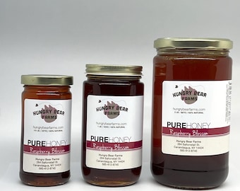May include: Three glass jars of honey with gold lids. The jars are labeled "Hungry Bear Farms" and "Pure Honey Raspberry Blossom". The jars are different sizes, with the largest jar being 2 lbs, the middle jar being 1 lb, and the smallest jar being 1/2 lb.
