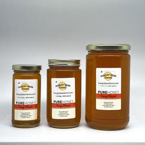 May include: Three glass jars of honey, labeled "Hungry Bear Farms", with the text "Pure Honey Orange Blossom" on the front. The jars are different sizes, with the largest jar being 2 pounds, the middle jar being 1 pound, and the smallest jar being 1/2 pound.