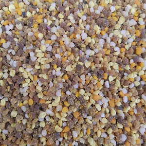 May include: Close-up of bee pollen granules in shades of brown, yellow, orange, and white. The granules are small and irregularly shaped, creating a textured surface. The image is well-lit, highlighting the natural colors and textures of the pollen.