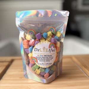 May include: A clear, iridescent bag filled with colorful, cube-shaped bath bombs. The bag features a label with the text "DeLYEtful" and "Rainbow Mini Bombs." The bath bombs are in various colors, including yellow, blue, purple, and orange.