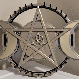 May include: A silver-coloured wall hanging featuring a pentagram, a triple moon symbol, and a central triquetra knot. The design is set against a circular backdrop with a black border and small, evenly spaced black accents. The piece is likely for spiritual or decorative purposes.
