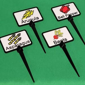 May include: Four garden plant markers with black stems and white rectangular labels. The labels display illustrations and names of vegetables: arugula, bell pepper, beets, and asparagus. The background is a solid green.