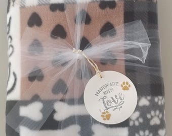 Puede incluir: Una manta de franela de retazos con una variedad de patrones con temática de perros, que incluyen huellas de patas, huesos, corazones y cuadros. La manta está atada con una cinta blanca y una etiqueta redonda que dice "Handmade with Love" con una huella de pata.
