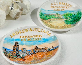Op de afbeelding: Twee witte keramische ring schaaltjes met handgeschilderde landschapsscènes en gouden letters. Eén schaaltje leest "ALI & MIKE ENGAGED 8.26.25 SEVILLE SPAIN". De andere leest "ANDREW & JULIANA ENGAGED 08.30.2025".
