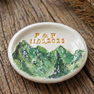 May include: A small, off-white ceramic ring dish with a hand-painted mountain scene in shades of green. The dish features the initials "P & F" and the date "11.05.2025" in gold lettering. The dish is round and sits on a wooden surface.