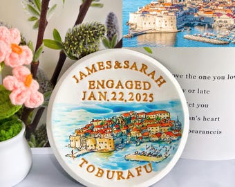 Op de afbeelding: Een witte keramische plaat met een handgeschilderde kustscène, met een stad met oranje daken en blauw water. De plaat is beschreven met "JAMES & SARAH ENGAGED JAN.22.2025" en "TOBURAFU" in gouden letters. Decoratieve bloemelementen zijn op de achtergrond.