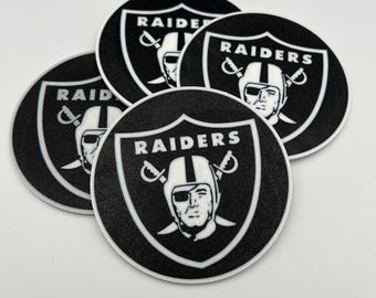 May include: Four black and white round coasters featuring the Raiders logo. Each coaster has a black background with a white border and the Raiders logo in the center. The logo includes the word "RAIDERS" and a graphic of a football player.