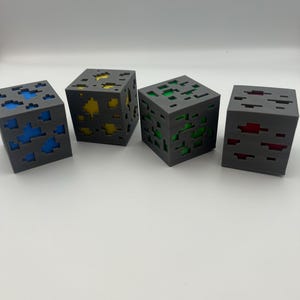 Könnte beinhalten: Vier graue, pixelige Würfel mit farbigen Innenräumen. Ein Würfel hat blau, ein anderer gelb, ein dritter grün und der letzte rot. Die Würfel sind mit einer blockartigen, geometrischen Ästhetik gestaltet, die an ein beliebtes Videospiel erinnert.