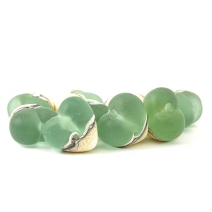 May include: A collection of sea glass heart-shaped beads in various shades of green. Each bead has a unique pattern of white and black swirls on a cream-colored base, creating a natural, organic aesthetic. The beads are arranged against a white background.