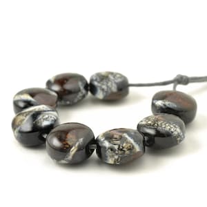 May include: A strand of eight dark brown and white beads. The beads are oval-shaped and strung on a gray cord. The beads have a glossy finish and a marbled appearance.