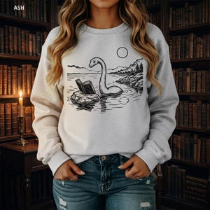 May include: A light gray crewneck sweatshirt featuring a black line art graphic of a long-necked creature resembling the Loch Ness Monster sitting on a rock reading an open book in a lake, with mountains and a circular moon in the background.