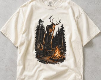 May include: An ivory short-sleeve t-shirt featuring a graphic print of a skeletal creature with large antlers standing by a campfire in a dark forest. The scene includes a small tent and tall pine trees. The shirt has a crew neckline and a label.