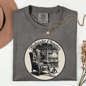 Op de afbeelding: Een antracietgrijs t-shirt met een ronde afbeelding van een skelet met een muts op, zittend in een fauteuil en een boek lezend in een bibliotheek. De tekst Midnight Chapter staat in een boog erboven. Een gouden ster-halsketting ligt op het t-shirt.