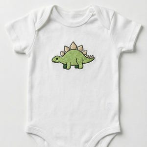 May include: White baby onesie with a green stegosaurus dinosaur embroidered on the front. The dinosaur has tan plates along its back and is outlined in black. The onesie has short sleeves and a round neckline.