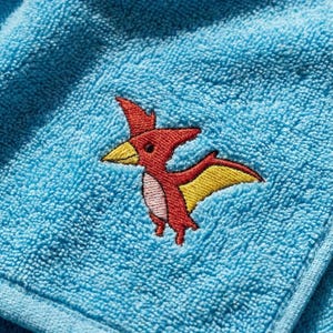 Op de afbeelding: Lichtblauw badstof handdoek met een geborduurd rood en geel pterodactylusontwerp. De pterodactylus heeft een gele snavel en vleugels en een roze buik. De handdoek is opgevouwen.