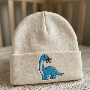 Puede incluir: Gorro de punto color crema con un diseño de dinosaurio azul bordado en la parte delantera. El dinosaurio tiene un cuello largo y hojas en la boca. El gorro tiene un borde doblado.