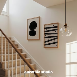 May include: Two framed abstract art prints with black shapes on a white background hang on a wall. One print features two overlapping black ovals, and the other has black stripes and dots. A staircase with a wooden handrail and a clear glass pendant light are also visible.