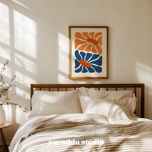 May include: A framed art print featuring two stylized flowers in orange and blue against a cream background. The artwork is displayed above a bed with striped bedding. The print is in a wooden frame.