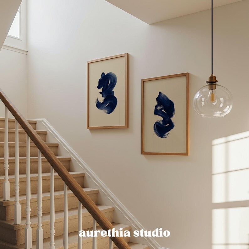May include: Two framed abstract art prints with navy blue brushstrokes on a cream background hang on a wall above a wooden staircase. The frames are light brown. A clear glass pendant light hangs from the ceiling. The text "aurethia studio" is at the bottom.
