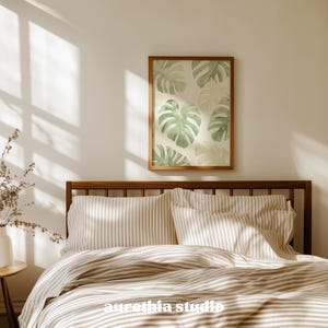 May include: A bedroom scene featuring a bed with striped bedding and a wooden headboard. Above the bed, a framed print displays watercolor-style monstera leaves in shades of green. The room is bathed in natural light, creating a warm and inviting atmosphere.